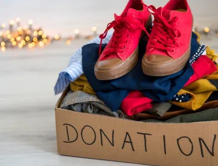 A cardboard donation box packed with folded clothes, a pair of red trainers on top